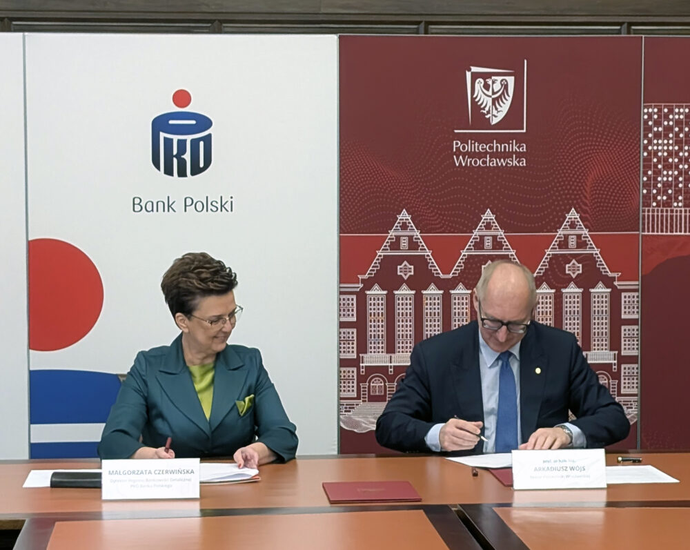 Małgorzata Czerwińska (Dyrektor Regionu Bankowości Detalicznej PKO Banku Polskiego) oraz prof. Arkadiusz Wójs (Rektor Politechniki Wrocławskiej) siedzą obok siebie przy dużym, drewnianym stole konferencyjnym i podpisują oficjalne dokumenty. Obydwie osoby uśmiechają się, patrząc na podpisywane papiery. Na stole przed nimi leżą tabliczki z ich imionami i nazwiskami. W tle znajdują się dwa duże banery: biały z logo PKO Bank Polski oraz bordowy z logo i grafiką Politechniki Wrocławskiej.