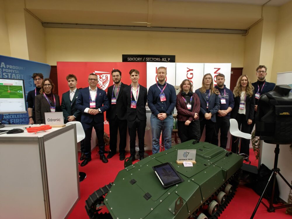 PWr na Made in Wroclaw 2026 - zdjęcie zbiorowe reprezentantów uczelni na stoisku, wśród zebranych prof. Piotr Młynarz, na pierwszym planie mały czołg